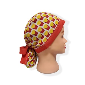 May include: Red and yellow patterned surgical scrub hat with a red tie. The hat is made of fabric and has a red trim.