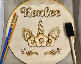 May include: A wooden craft kit featuring a unicorn design with the name "Kenlee" painted on it. The kit includes six small paint pots in various colors, two paintbrushes, a sponge brush, and a wooden round base.