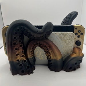May include: A 3D printed octopus stand for a Nintendo Switch console. The stand is black and gold with a textured octopus design. The Switch console is in the stand and the screen is facing forward.
