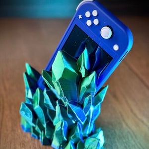 Może przedstawiać: Niebieska konsola Nintendo Switch Lite jest wyświetlana na stojaku wydrukowanym w 3D z zielonych i niebieskich kryształów.