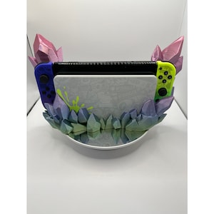 May include: A white Nintendo Switch console with a green and white design is displayed in a crystal-shaped stand. The stand is made of iridescent plastic and features a variety of colours, including purple, blue, and green. The console is accompanied by two controllers, one blue and one green.