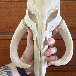 Peut inclure: R&eacute;plique de casque cr&acirc;ne Mandalorien de couleur cr&egrave;me. Le casque pr&eacute;sente un design d&eacute;taill&eacute; avec des d&eacute;fenses et des orbites pro&eacute;minentes. La sculpture est tenue dans une main, mettant en valeur sa taille et sa texture. L'esth&eacute;tique g&eacute;n&eacute;rale sugg&egrave;re des objets de collection de science-fiction ou fantastiques.