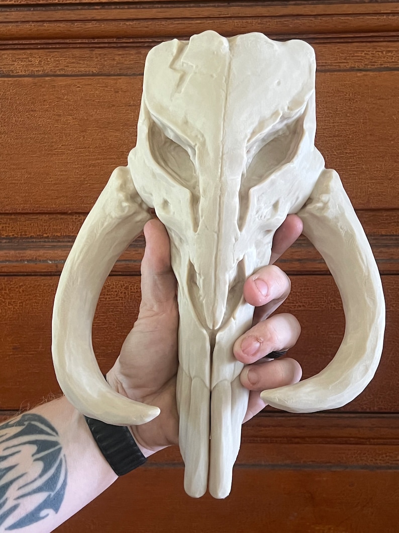 Peut inclure: R&eacute;plique de casque cr&acirc;ne Mandalorien de couleur cr&egrave;me. Le casque pr&eacute;sente un design d&eacute;taill&eacute; avec des d&eacute;fenses et des orbites pro&eacute;minentes. La sculpture est tenue dans une main, mettant en valeur sa taille et sa texture. L'esth&eacute;tique g&eacute;n&eacute;rale sugg&egrave;re des objets de collection de science-fiction ou fantastiques.