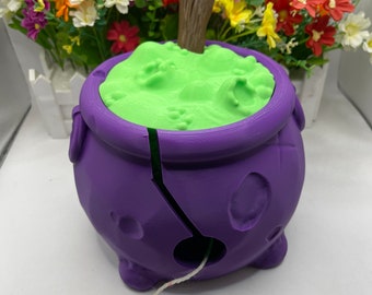 May include: Purple cauldron yarn bowl with a green lid and a wooden spoon. The bowl has a hole in the side for yarn to feed through. White yarn is wrapped around the bowl.