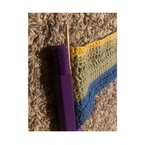 Puede incluir: Primer plano de un proyecto de ganchillo en progreso, con un patrón de rayas multicolores de amarillo, verde y azul. Se puede ver un gancho de ganchillo morado en la imagen.