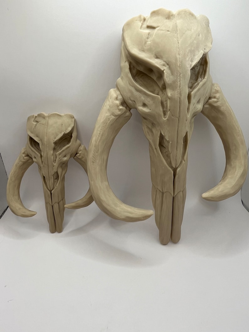 Peut inclure: Deux sculptures de cr&acirc;nes stylis&eacute;s, de couleur blanc cass&eacute;, avec des cornes incurv&eacute;es. Le plus grand cr&acirc;ne est dress&eacute;, tandis qu'une version plus petite est &agrave; gauche. Les deux ont des traits faciaux d&eacute;taill&eacute;s et une surface textur&eacute;e.
