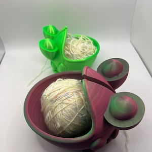 May include: Two 3D printed yarn bowls, one green and one with a pink and green ombre finish. Both bowls have a unique design and are holding balls of yarn.