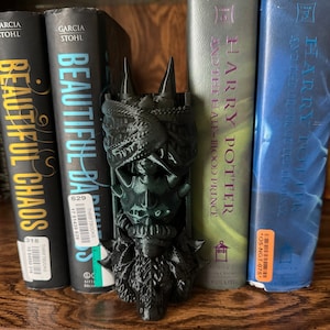 May include: A black and green 3D printed bookend shaped like a dragon's head with a textured surface. The bookend is holding up two books titled "Beautiful Chaos" and "Beautiful Darkness".