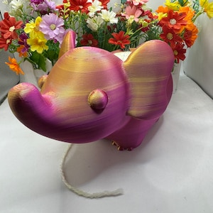 Puede incluir: Una figurita de elefante rosa y dorada con una cola blanca. El elefante está hecho de un material texturizado y tiene un aspecto caprichoso.