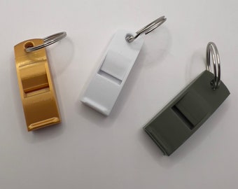 Op de afbeelding: Drie sleutelhangers met fluitjes. Eén is goud, één is wit en één is donkergrijs.