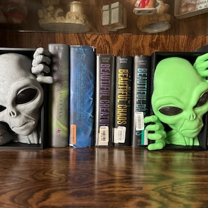 Peut inclure: Paire de serre-livres sur le thème des extraterrestres, gris et vert. Le serre-livres gris représente une tête d'extraterrestre avec des mains, tandis que le serre-livres vert a un design similaire. Des livres avec des titres comme "Dune" sont visibles.
