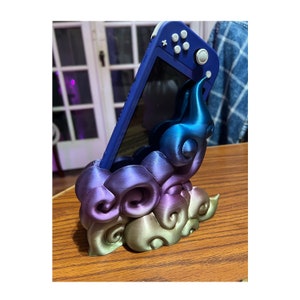 May include: A blue and purple 3D printed stand for a Nintendo Switch console. The stand is shaped like a cloud with a swirl design. The Switch console is in the stand and is facing the camera.