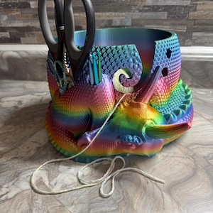 May include: A rainbow-colored unicorn-shaped desk organizer. The organizer holds scissors, pens, and other small items. The design features a textured, scaly pattern and a curled horn. A white string is draped across the front.