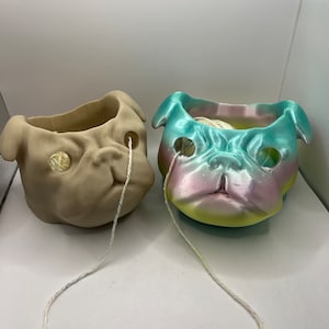 May include: Two ceramic pug-shaped yarn bowls, one in a natural beige colour and the other in a multi-coloured ombre glaze. Both bowls have yarn coming out of their mouths.