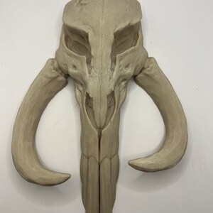 Peut inclure: Un cr&acirc;ne stylis&eacute; de couleur beige avec des cornes incurv&eacute;es, rappelant un casque Mandalorien. La sculpture pr&eacute;sente une surface textur&eacute;e et d&eacute;taill&eacute;e et est probablement un objet d&eacute;coratif ou une d&eacute;coration murale.