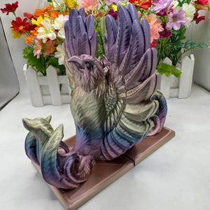 Peut inclure: Un serre-livres multicolore représentant un phénix avec ses ailes déployées. Le serre-livres est fait d'un matériau imprimé en 3D et présente un dégradé de couleurs allant du violet au bleu et au vert.