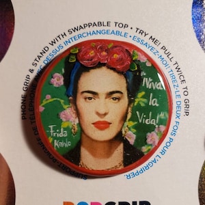May include: A circular phone grip featuring a portrait of Frida Kahlo with red flowers in her hair, green background, and the text Viva la Vida and Frida Kahlo. The packaging includes the text Phone Grip & Stand with Swappable Top and PopGrip.