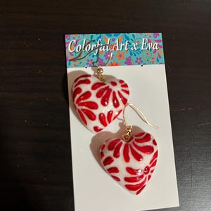 May include: A pair of heart-shaped earrings with a white base and a red, swirling design. The earrings are on gold-colored hooks and are displayed on a white card with the text "Colorful Art x Eva".