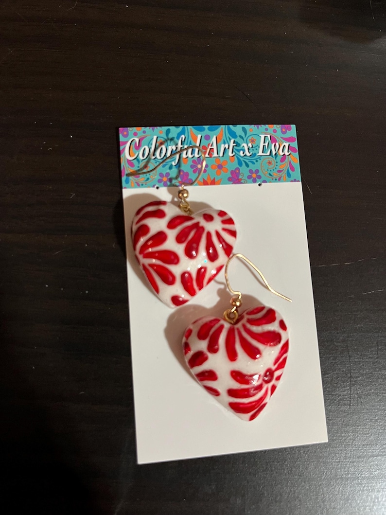 May include: A pair of heart-shaped earrings with a white base and a red, swirling design. The earrings are on gold-colored hooks and are displayed on a white card with the text "Colorful Art x Eva".