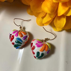 May include: A pair of heart-shaped earrings with a white base, adorned with colorful, abstract designs in red, orange, green, blue, and purple. The earrings have gold-colored hooks and are set against a blurred background.