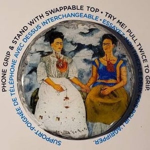 May include: A round, white phone grip with a colorful, holographic design. The grip features a picture of two women with painted hearts on their chests. The text "TRY ME! PULL TWICE TO GRIP" is printed around the edge of the grip.