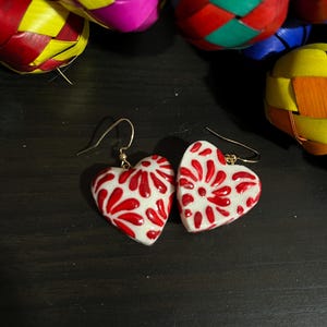 May include: A pair of heart-shaped earrings with a white base and a red floral design. The earrings have gold-colored hooks. Colorful woven ornaments are in the background.