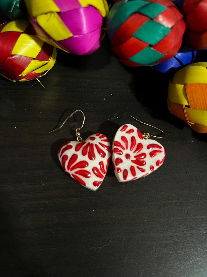 May include: A pair of heart-shaped earrings with a white base and a red floral design. The earrings have gold-colored hooks. Colorful woven ornaments are in the background.