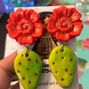 May include: A pair of handmade polymer clay earrings featuring a red flower with a yellow center and a green prickly pear cactus. The earrings are on a white card with the text "colorfulartxeva" on it.