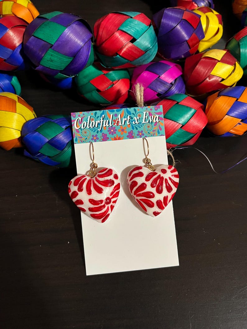 May include: White heart-shaped earrings with red floral designs. The earrings are hanging on a white card with the text "Colorful Art x Eva". Colorful woven spheres in various colors are in the background.