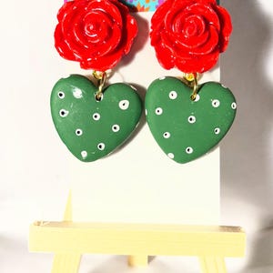 May include: A pair of handmade earrings featuring red rose tops and green heart-shaped pendants with white polka dots. The earrings hang from gold-colored hooks. The background includes a sign with the text "Colorful Art x Eva".