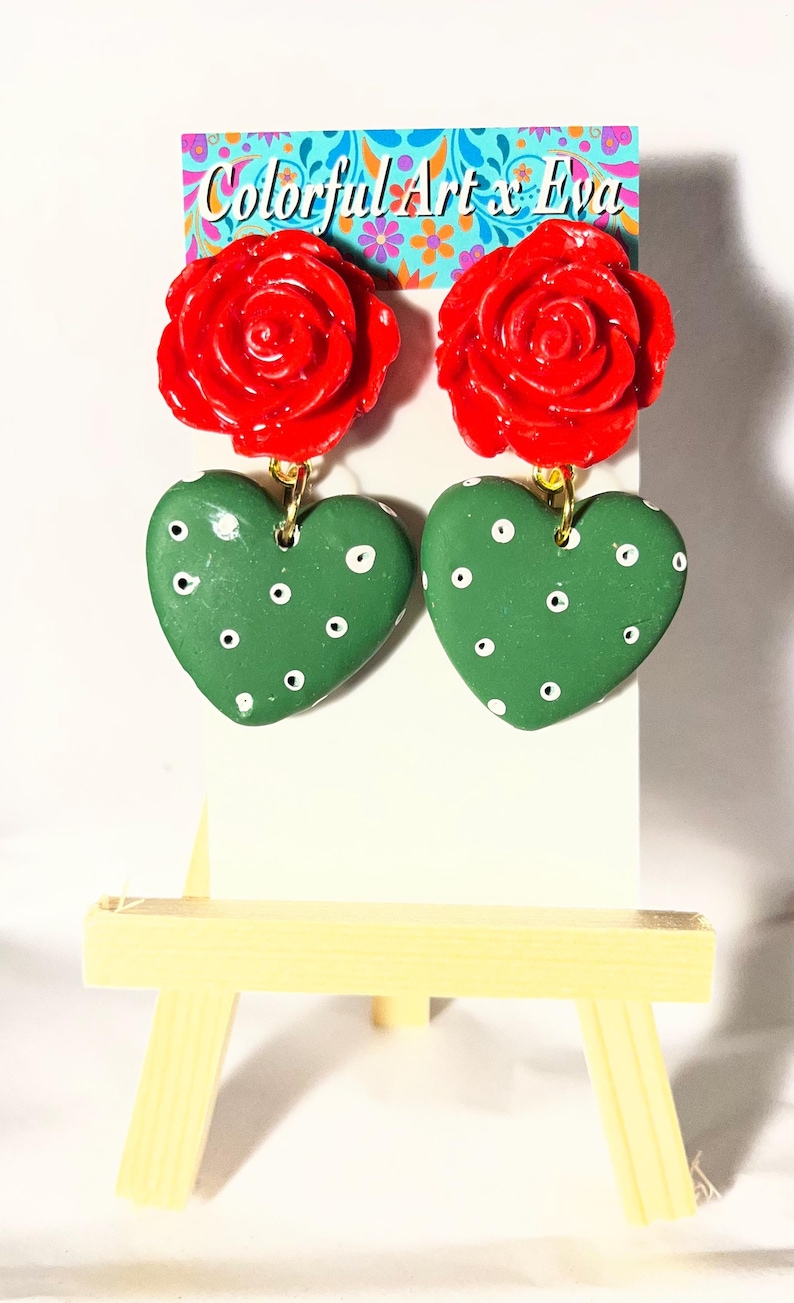 May include: A pair of handmade earrings featuring red rose tops and green heart-shaped pendants with white polka dots. The earrings hang from gold-colored hooks. The background includes a sign with the text "Colorful Art x Eva".