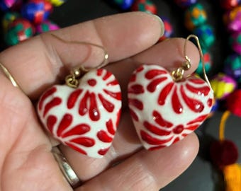 May include: A pair of heart-shaped earrings featuring a white ceramic base with a glossy red floral pattern. The earrings are attached to gold-toned metal hooks, showcasing a smooth, rounded surface with a distinct hand-painted aesthetic.