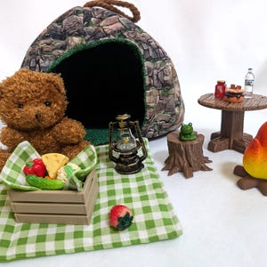 Puede incluir: Un oso de peluche marrón está sentado en una manta a cuadros verde y blanca con una caja de madera de comida. Una escena de campamento en miniatura incluye una tienda de campaña, una linterna, una mesa con comida, una fogata y un tocón de árbol con una rana.