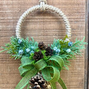 Puede incluir: Una corona r&uacute;stica hecha de cuerda con un lazo de arpillera verde y pi&ntilde;as. La corona est&aacute; decorada con ramitas de vegetaci&oacute;n y peque&ntilde;as bayas azules.