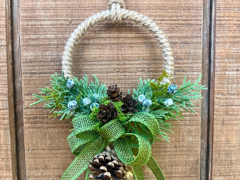 Puede incluir: Una corona r&uacute;stica hecha de cuerda con un lazo de arpillera verde y pi&ntilde;as. La corona est&aacute; decorada con ramitas de vegetaci&oacute;n y peque&ntilde;as bayas azules.