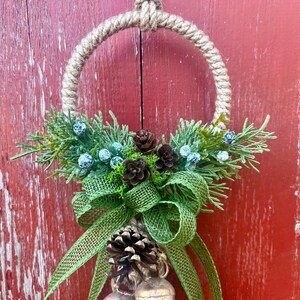 Puede incluir: Una corona de Navidad r&uacute;stica con un c&iacute;rculo de cuerda de arpillera, ramas de pino verdes, bayas azules, pi&ntilde;as marrones y un lazo de arpillera verde. La corona est&aacute; colgada en una puerta de madera roja.