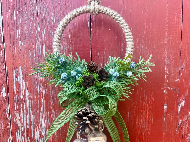 Puede incluir: Una corona de Navidad r&uacute;stica con un c&iacute;rculo de cuerda de arpillera, ramas de pino verdes, bayas azules, pi&ntilde;as marrones y un lazo de arpillera verde. La corona est&aacute; colgada en una puerta de madera roja.