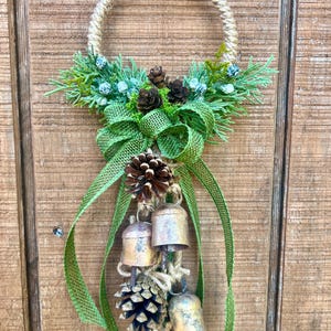 Puede incluir: Una corona r&uacute;stica hecha de cuerda con un lazo de arpillera verde y pi&ntilde;as. La corona est&aacute; decorada con peque&ntilde;as campanas y vegetaci&oacute;n.