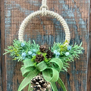 Puede incluir: Una corona r&uacute;stica hecha de cuerda natural con un lazo de arpillera verde y pi&ntilde;as. La corona est&aacute; decorada con ramitas de vegetaci&oacute;n y peque&ntilde;as bayas azules.