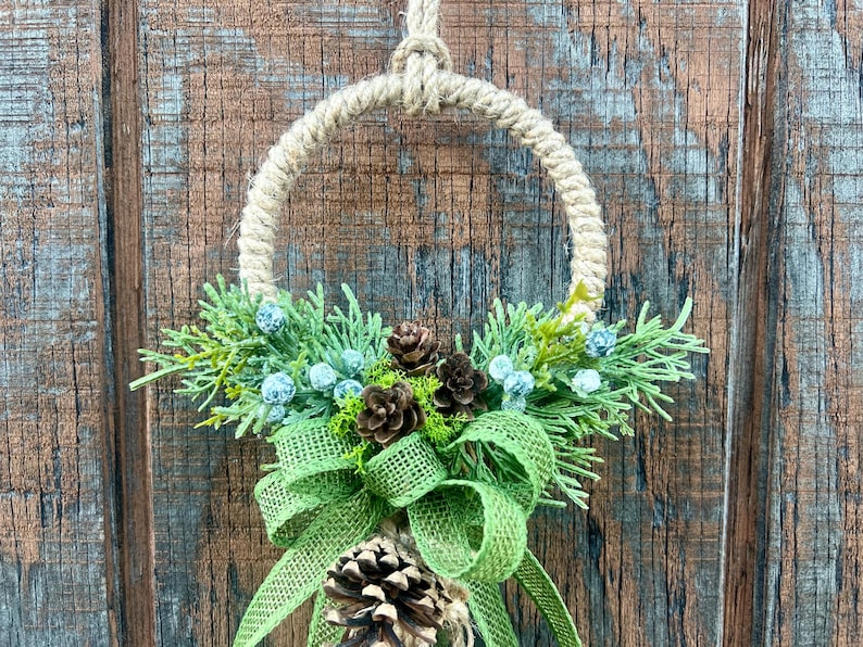 Puede incluir: Una corona r&uacute;stica hecha de cuerda natural con un lazo de arpillera verde y pi&ntilde;as. La corona est&aacute; decorada con ramitas de vegetaci&oacute;n y peque&ntilde;as bayas azules.