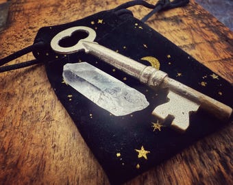 Llave antigua con forma de esqueleto y punta de cristal de cuarzo transparente: amuleto de oportunidad y protección, en una bolsita de terciopelo negro con luna creciente y estrellas.