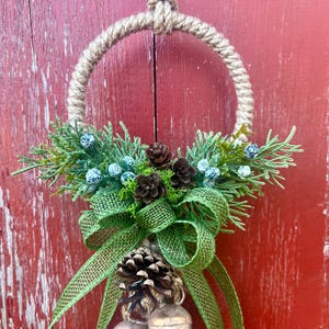 Puede incluir: Una corona de Navidad r&uacute;stica con un c&iacute;rculo de cuerda de arpillera, ramas de pino verdes, bayas azules, pi&ntilde;as marrones y un lazo de arpillera verde. La corona est&aacute; colgada en una puerta de madera roja.