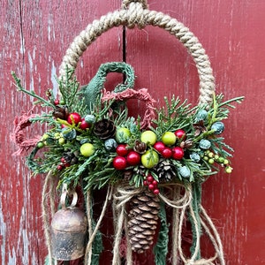 May include: A rustic Christmas wreath featuring a brown pine cone, red berries, and green foliage. The wreath is made with twine and has a bell hanging from it.