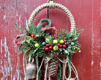 May include: A rustic Christmas wreath featuring a brown pine cone, red berries, and green foliage. The wreath is made with twine and has a bell hanging from it.