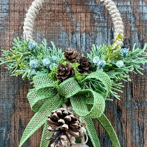 Puede incluir: Una corona de Navidad r&uacute;stica con un lazo de arpillera, pi&ntilde;as y vegetaci&oacute;n. Dos peque&ntilde;as campanas cuelgan de la parte inferior de la corona.