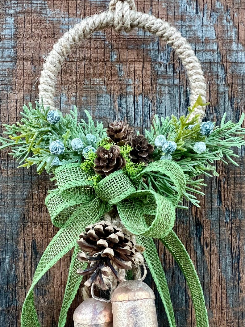 Puede incluir: Una corona de Navidad r&uacute;stica con un lazo de arpillera, pi&ntilde;as y vegetaci&oacute;n. Dos peque&ntilde;as campanas cuelgan de la parte inferior de la corona.