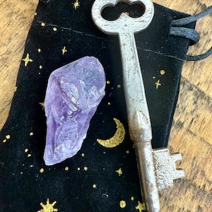 May include: An amethyst crystal and a vintage metal key rest on a black velvet pouch. The pouch is decorated with gold stars and a crescent moon. The key has a worn, antique appearance.