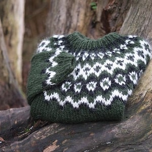 May include: A green, white, and black knitted sweater with a traditional Scandinavian pattern. The sweater is folded and placed on a tree trunk.