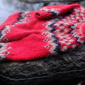 Könnte beinhalten: Ein roter, grauer und blauer Strickpullover mit traditionellem skandinavischem Muster. Der Pullover ist gefaltet und liegt auf einem grauen Stein.