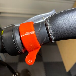 May include: Orange handlebar grip with a lever on the side. The grip is attached to a black handlebar with a black leather cover.